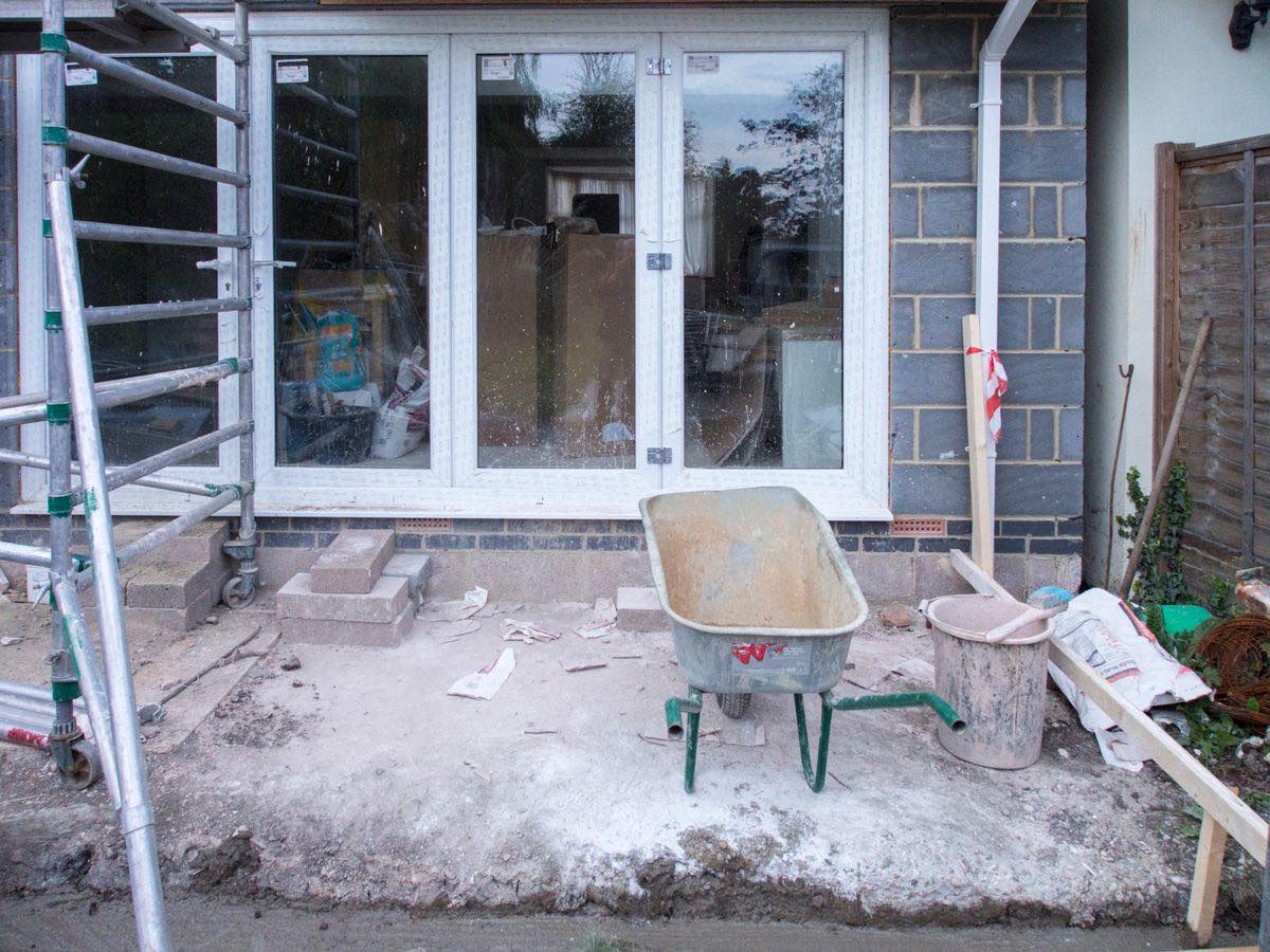 The back of a house with an extension under construction: new glazed doors set in block walls, a scaffold tower, a wheelbarrow and a bucket on a bare concrete slab.