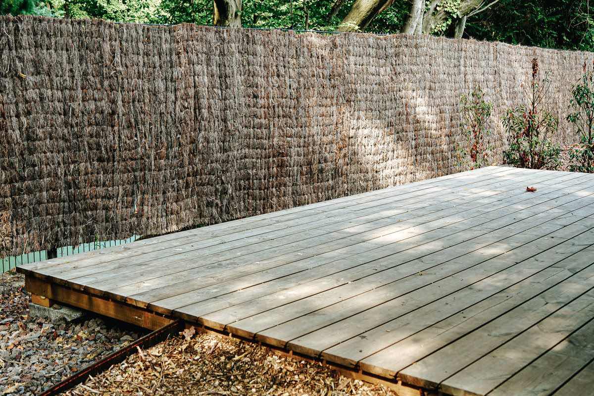 A low timber deck of long boards laid side by side, with its framing visible along the front edge, in front of a screened fence.