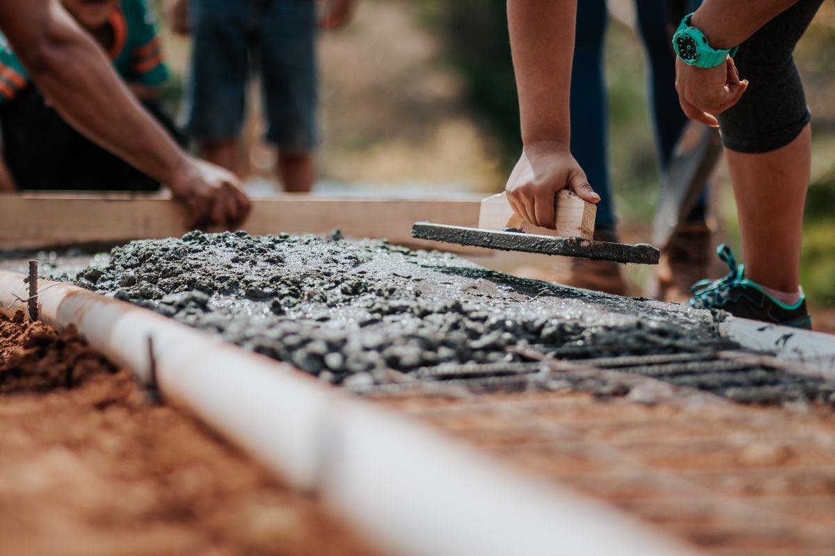 Fresh concrete being spread and levelled inside timber boxing over reinforcing mesh, with workers' hands and legs in view.
