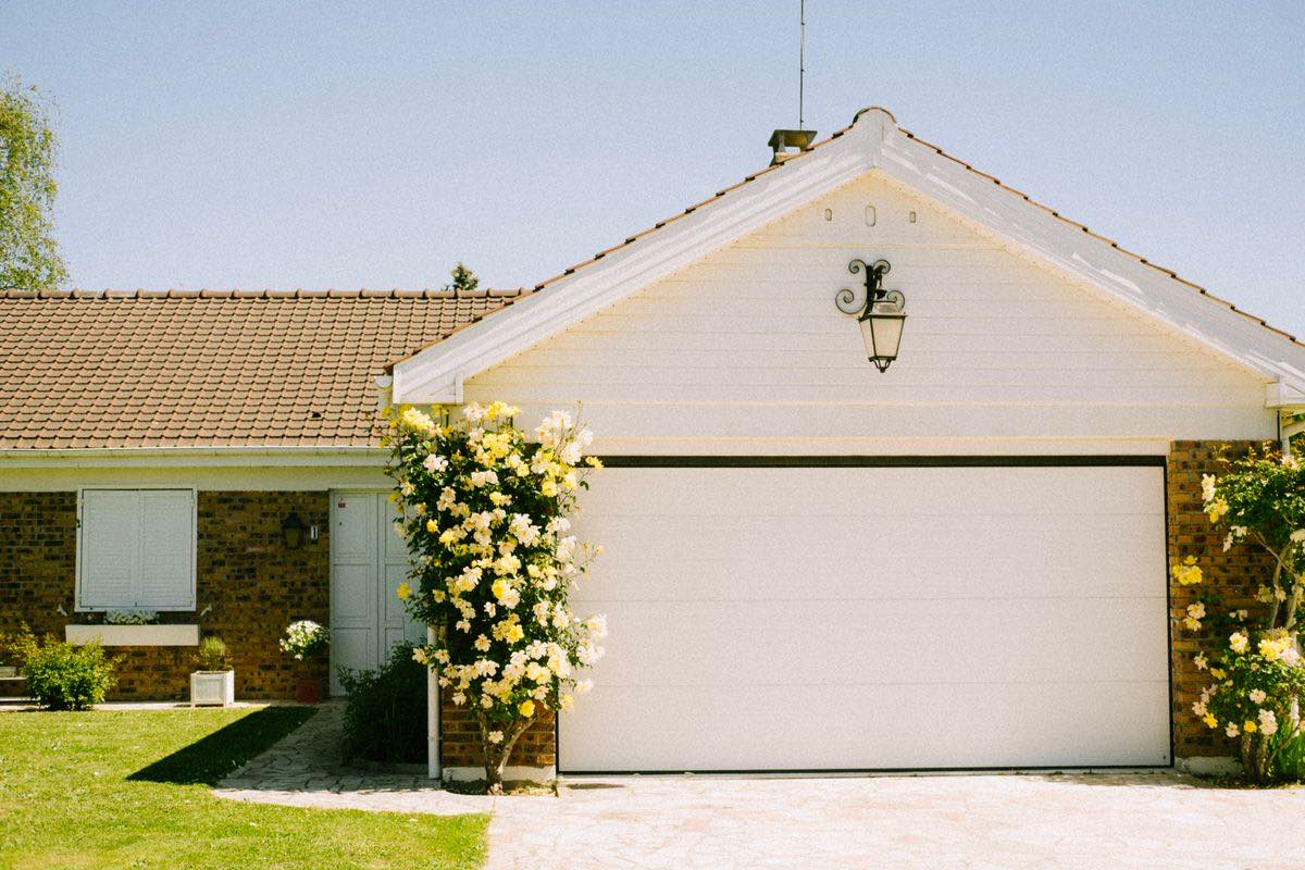 The front of a house with a closed white garage door under a gable, a lamp on the wall and yellow roses beside it.