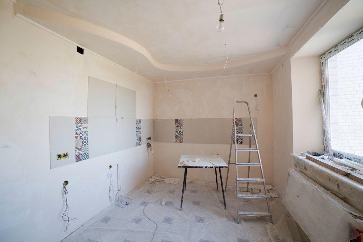 A bare kitchen room under renovation, with plastered walls, a few tiles stuck up on the wall, a stepladder, a table and plastic sheeting on the floor.