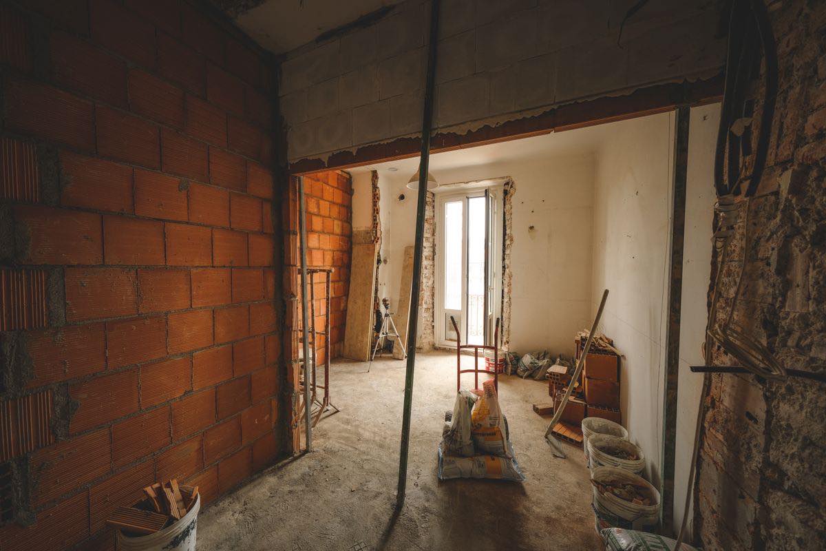 A room stripped back to brick and bare floor with a prop holding up a new opening, bags of material, buckets, a tripod and a door to a balcony in the far wall.